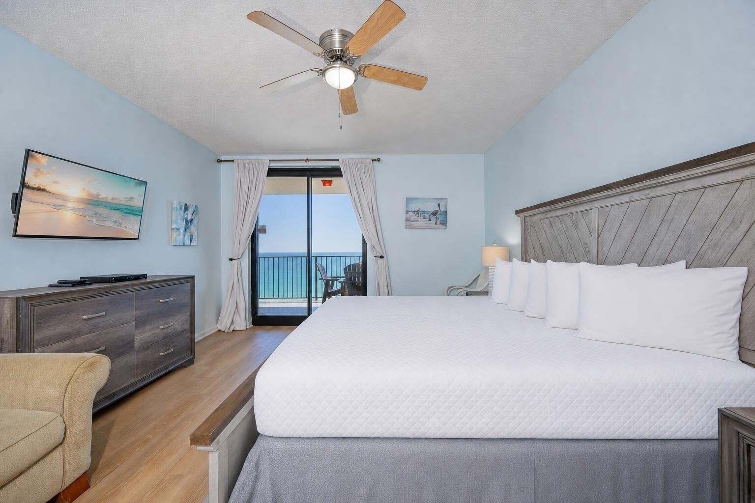 A hotel room with a bed, a ceiling fan, a TV, a dresser, and a view of the ocean.