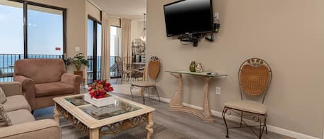A living room with a sofa, armchair, and a glass coffee table. There is a television mounted on the wall and a dining area in the background.
