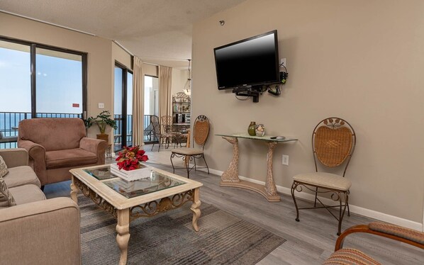 Una sala de estar con un sofá, un sillón y una mesa de centro de vidrio. Hay un televisor montado en la pared y un comedor al fondo.