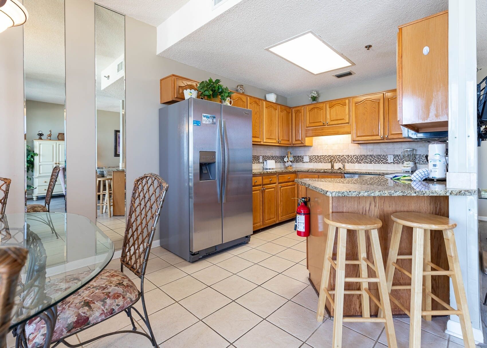 A modern kitchen with wooden cabinets, a stainless steel refrigerator, a granite countertop, and a glass dining table with chairs.