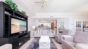 A living room with a flat-screen TV, a ceiling fan, a sofa, a coffee table, and a kitchen in the background.