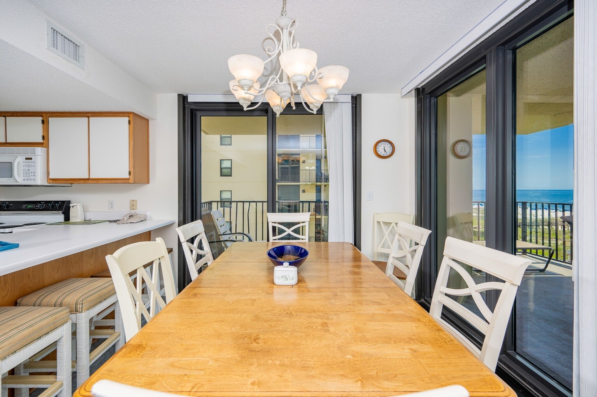 A dining area with a wooden table, white chairs, a chandelier, and a view of the sea through large windows.