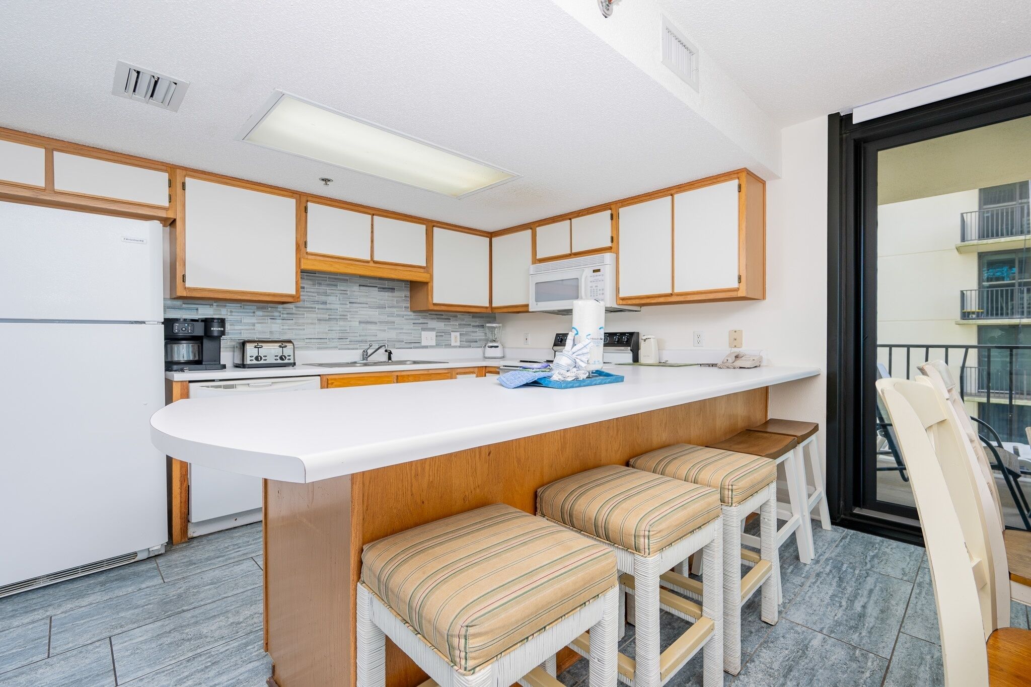 A modern kitchen with white cabinets, a central island with bar stools, and a view of the balcony through the glass door.