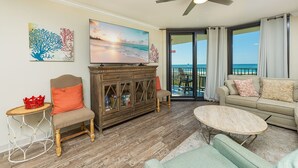 A living room with a television, a wooden cabinet, a sofa, a coffee table, and a view of the ocean through a sliding door. - Phoenix Vii 7316 2 Bedroom Condo (Orange Beach)