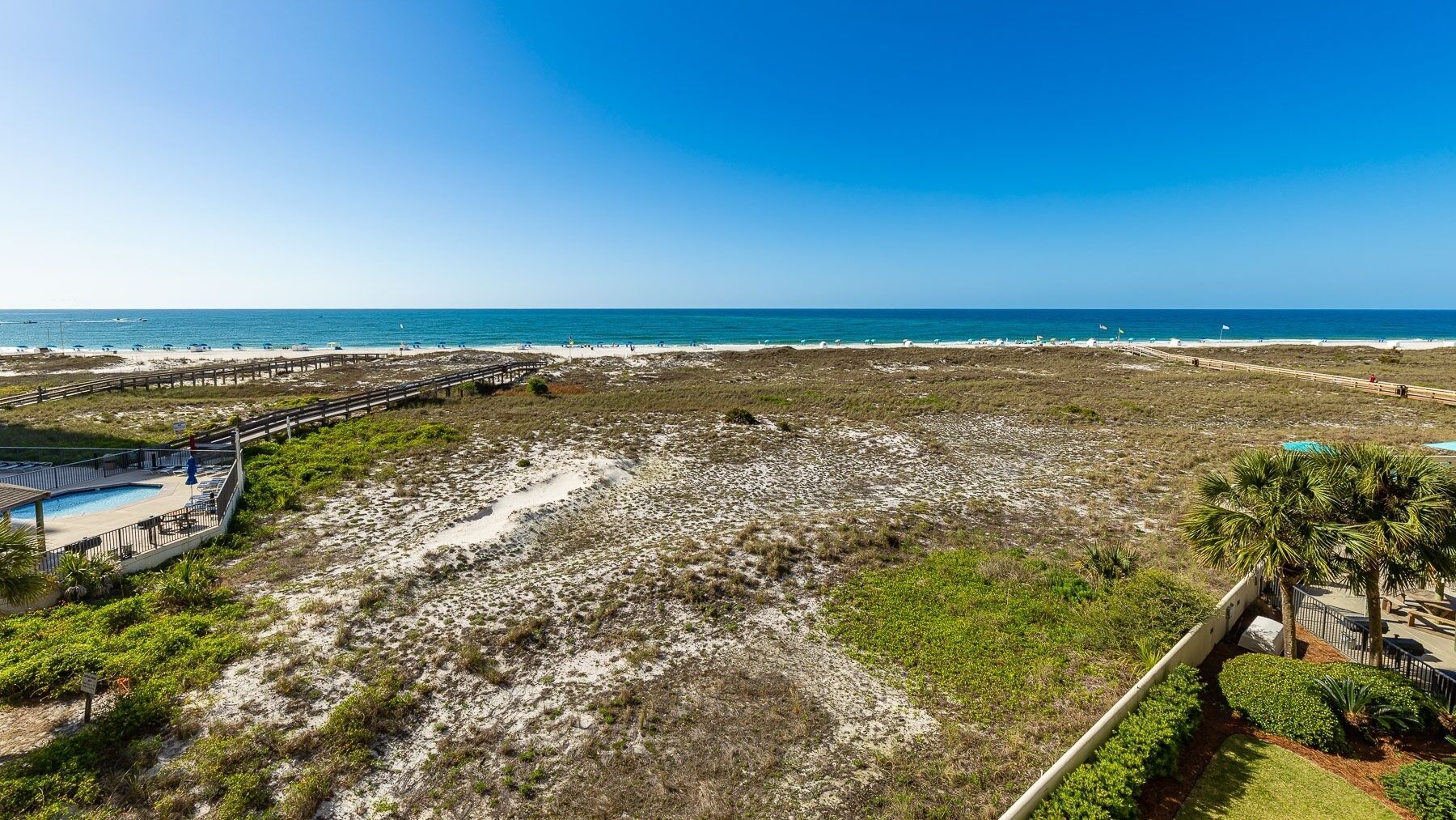 Blick auf einen Strand mit einem Holzsteg, einem Poolbereich und blauem Himmel.