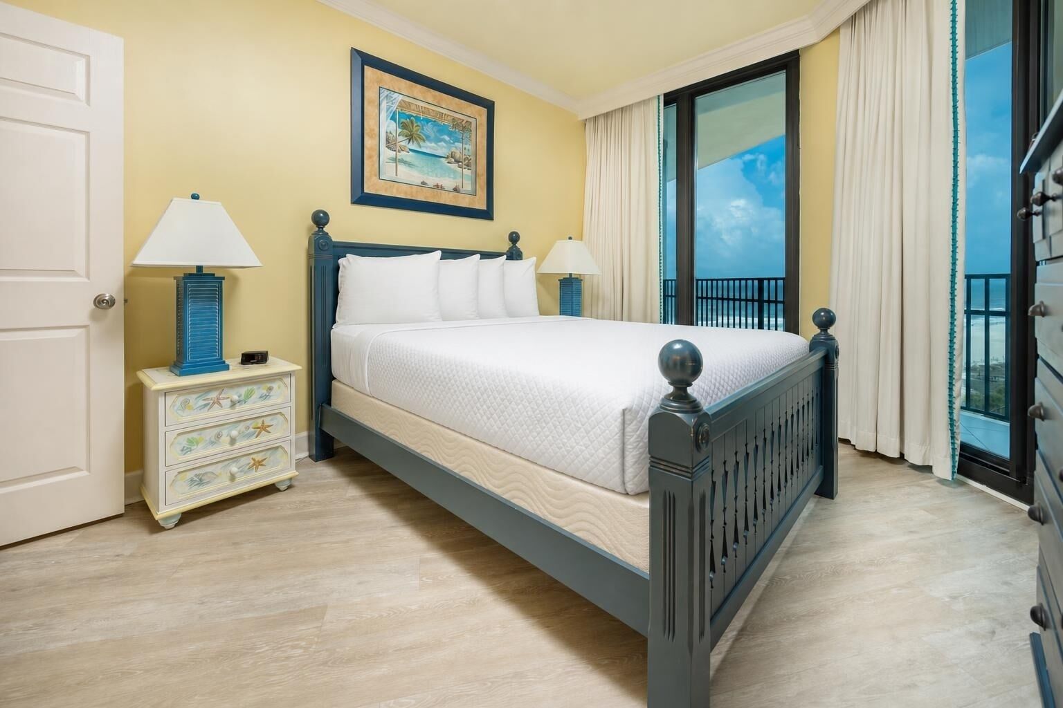 A bed with a headboard, a nightstand with a lamp, a dresser, a painting on the wall, and a view through a window.