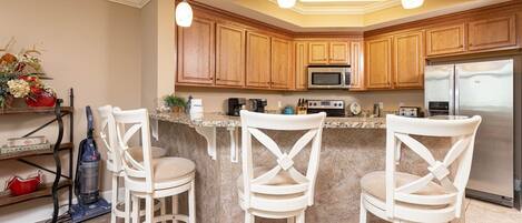 A kitchen with wooden cabinets, a microwave, a refrigerator, and three white chairs.