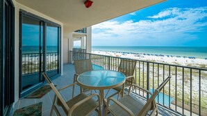 A balcony with a glass table and chairs, overlooking a beach with umbrellas and a clear sky.
