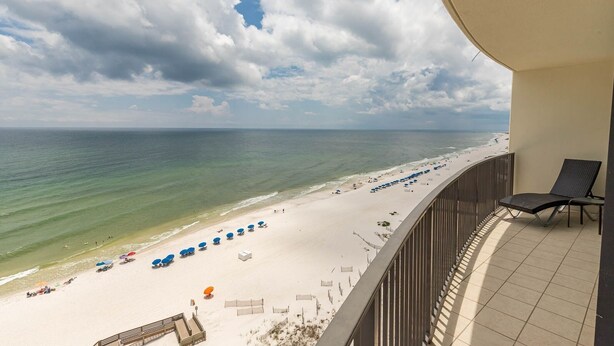 Blick auf einen Strand vom Balkon aus, mit Geländer, einem Stuhl und Strandsonnenschirmen.