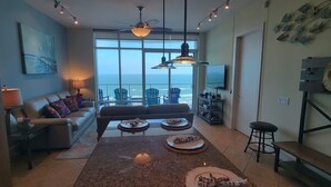 A living room with a sofa, coffee table, and chairs, a dining area with a table and chairs, and a large window with a view of the ocean. - Sapphire 1706 3 Bedroom Home by RedAwning (South Padre Island)