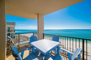 Un balcon avec une table blanche et des chaises bleues, donnant sur une plage et l’océan.