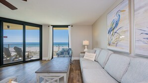 A living room with a sofa, a table, and a chair, a balcony with a view of the beach, and a painting on the wall. - Phoenix Iv 4023 3 Bedroom Condo (Orange Beach)