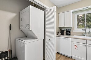 A compact kitchen with a built-in washer and dryer, a coffee maker, and a window with a view of greenery. - 5d Amelia Landings 2 Bedroom Condo by RedAwning (Fernandina Beach)