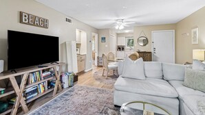 A living room with a TV, bookshelves, a white sofa, and a dining area with chairs and a table. - 5d Amelia Landings 2 Bedroom Condo by RedAwning (Fernandina Beach)
