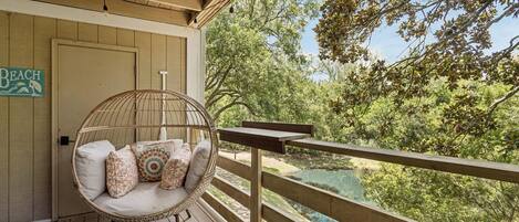 A wooden deck with a wicker chair and cushions, overlooking a natural landscape with trees and a body of water.