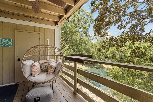 A wooden deck with a wicker chair and cushions, overlooking a natural landscape with trees and a body of water.