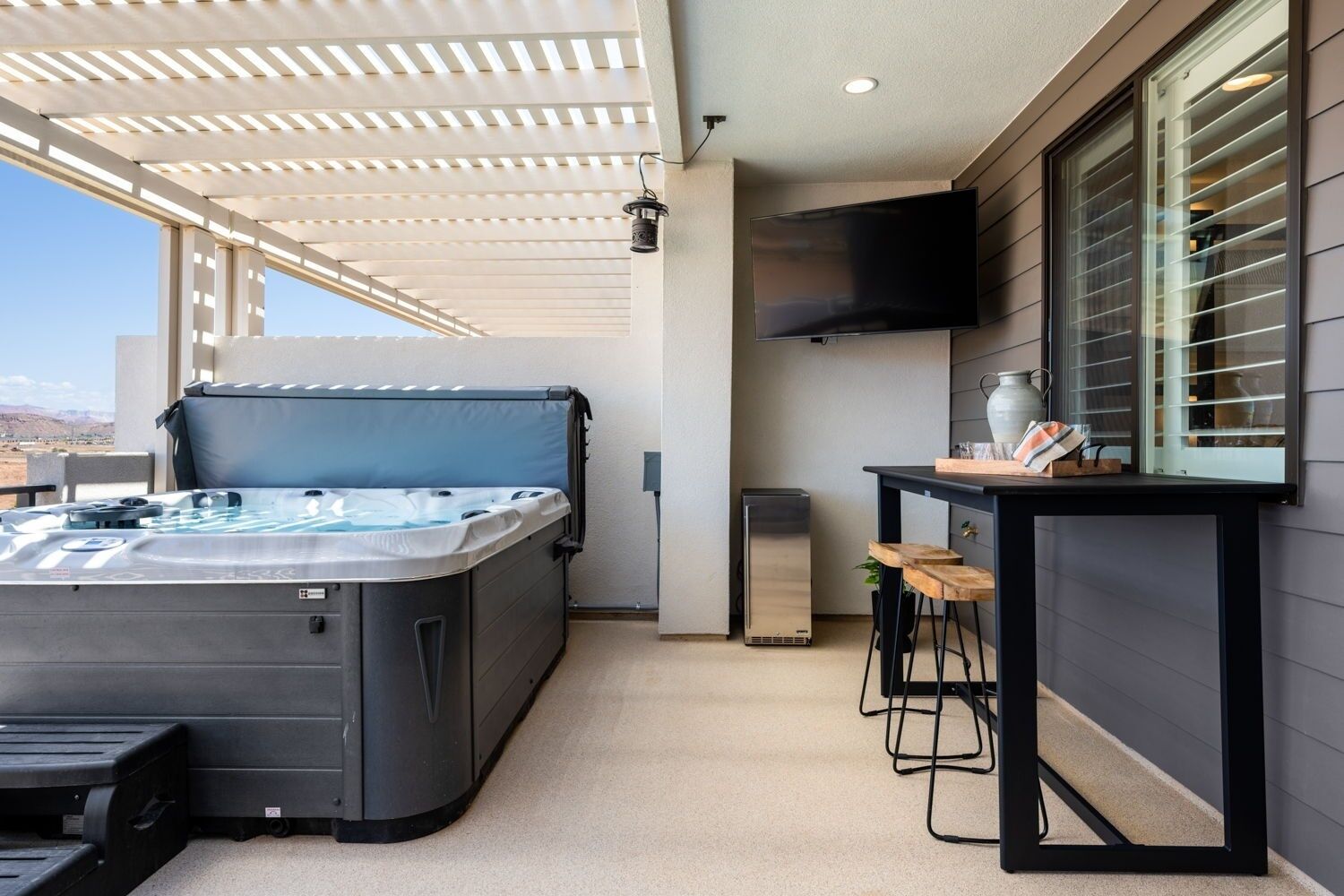Un jacuzzi installé sous une pergola, un téléviseur fixé au mur et un coin bar avec des tabourets.