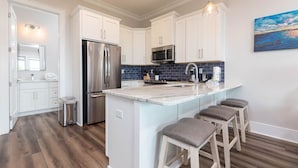 A modern kitchen with white cabinets, a stainless steel refrigerator, a microwave, and a blue tiled backsplash. There are three bar stools and a painting on the wall. - West Side Cottages N 3 Bedroom Home (Gulf Shores)