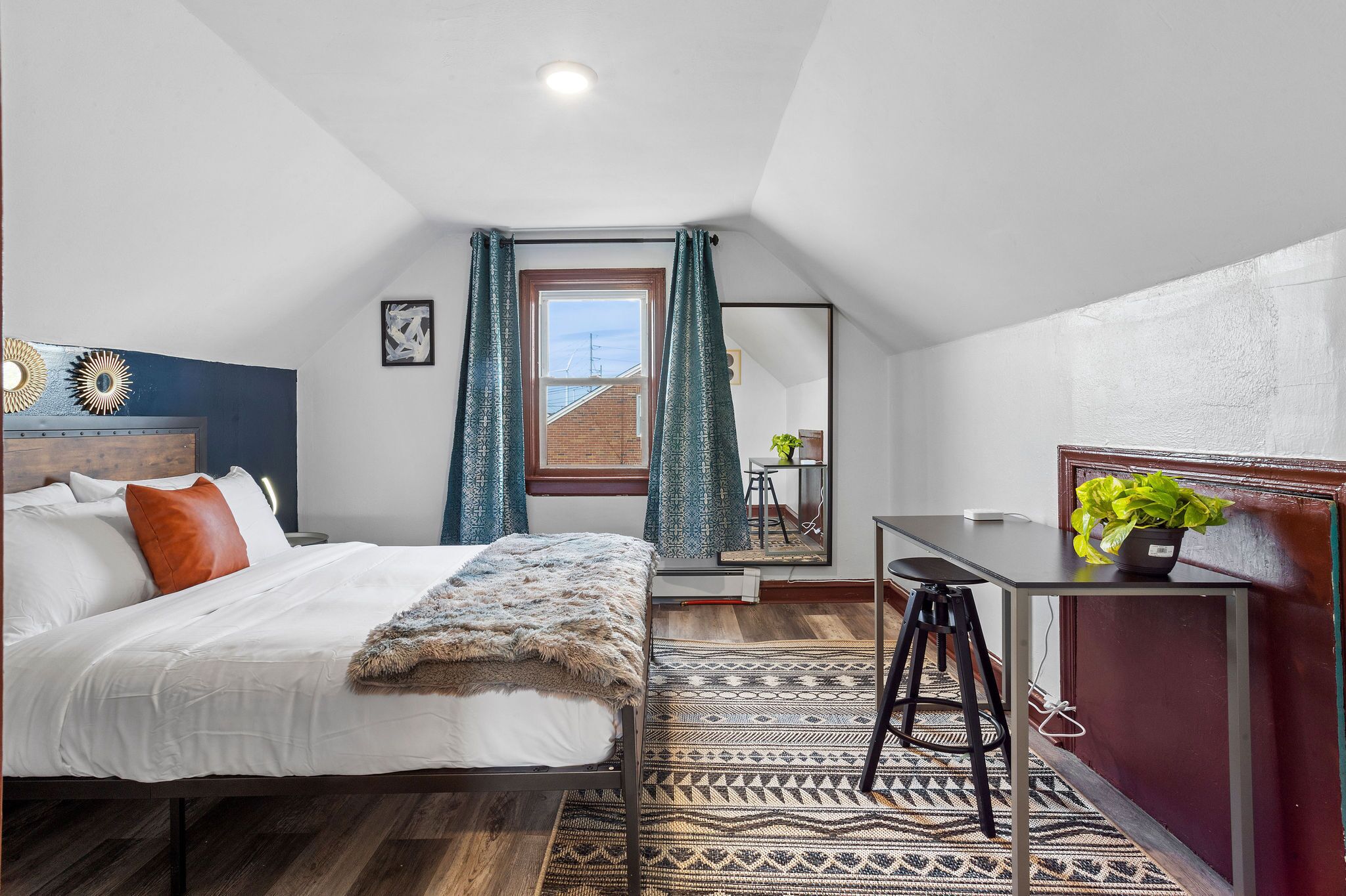 A bedroom with a bed, a desk, a chair, a mirror, and a window with curtains.