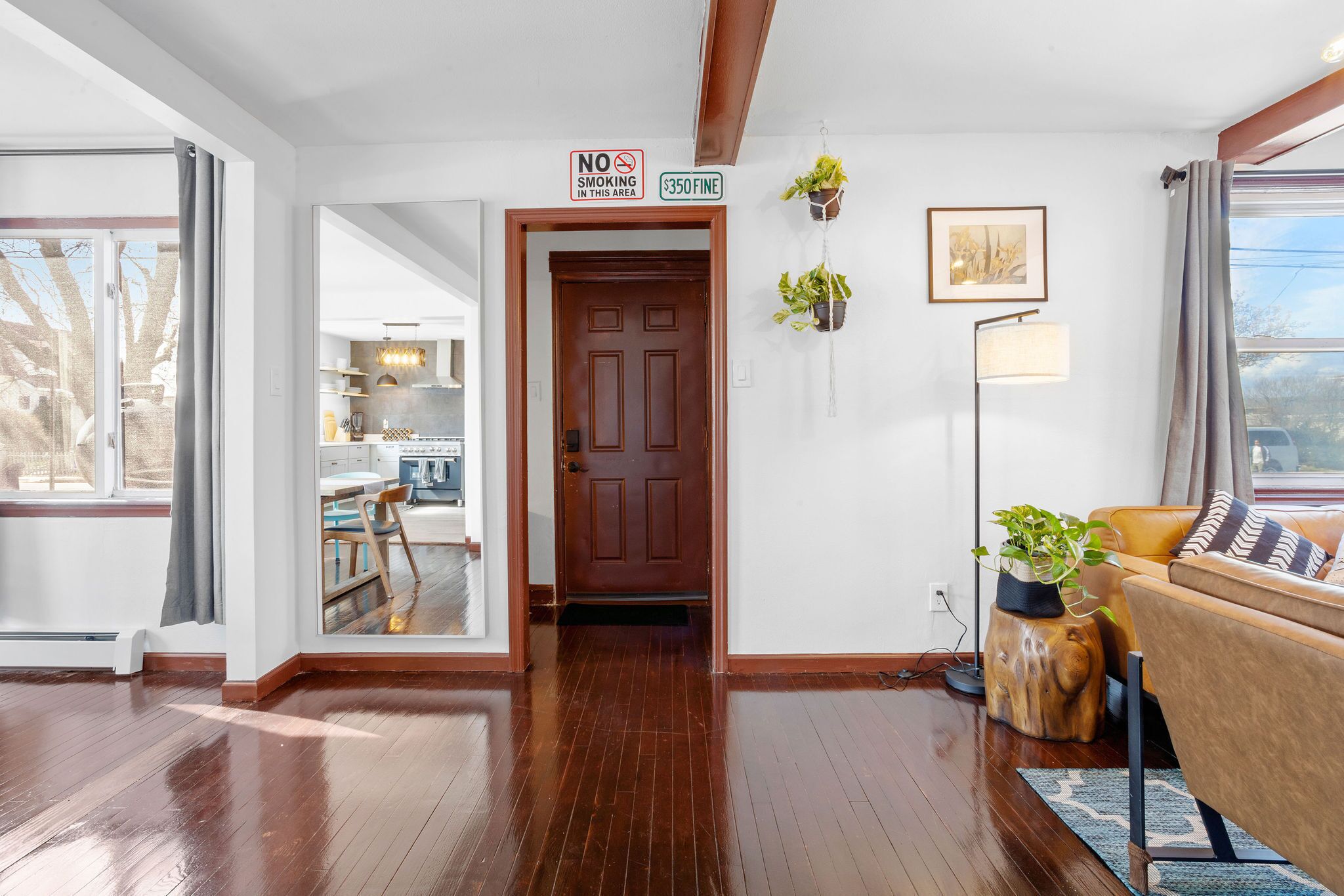 A hallway with a wooden floor, a dining area with a table and chairs, a kitchen in the background, and a living area with a sofa and plants.
