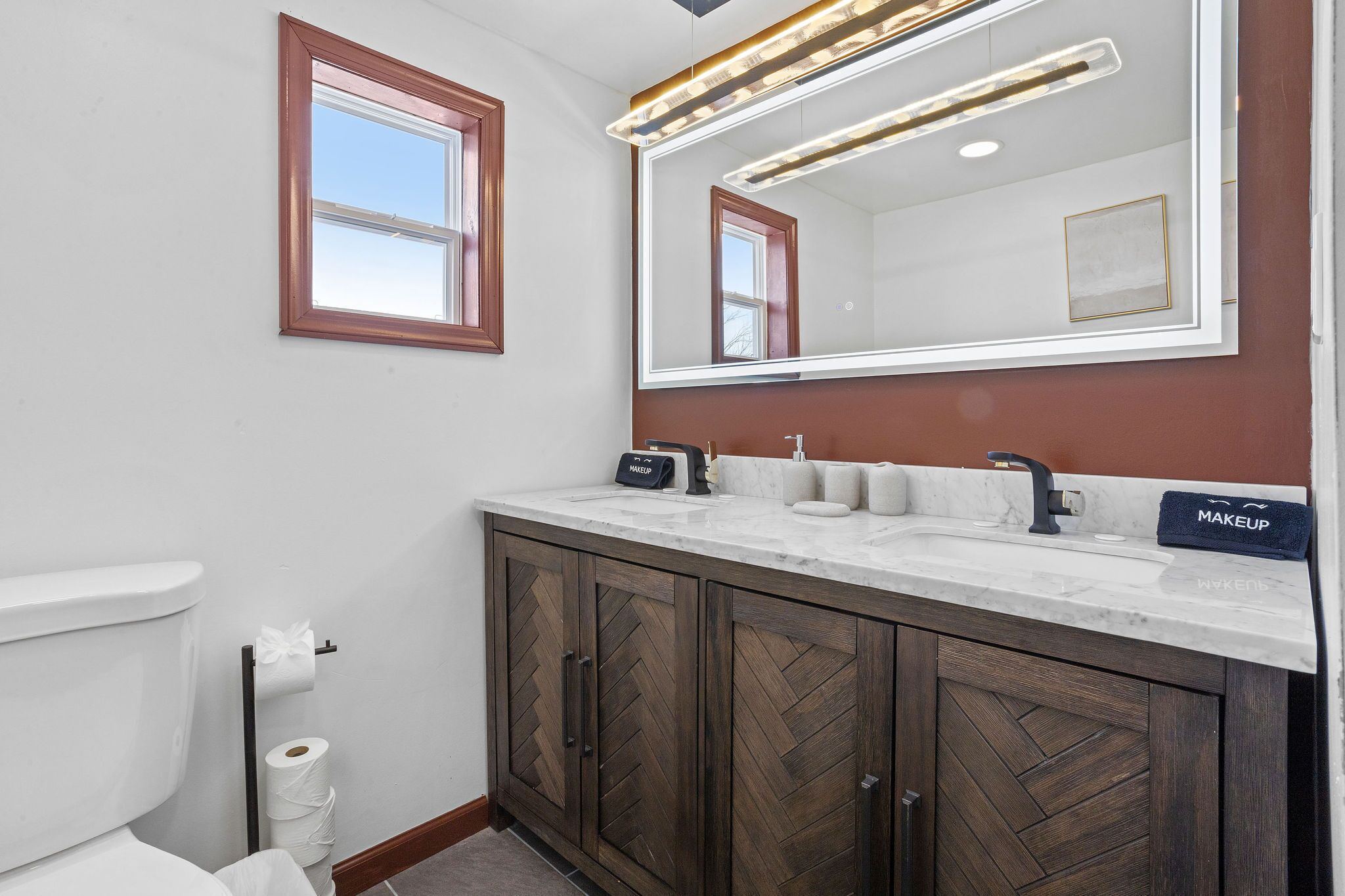 A bathroom with a white toilet, a wooden herringbone cabinet, a large mirror, and a modern light fixture.