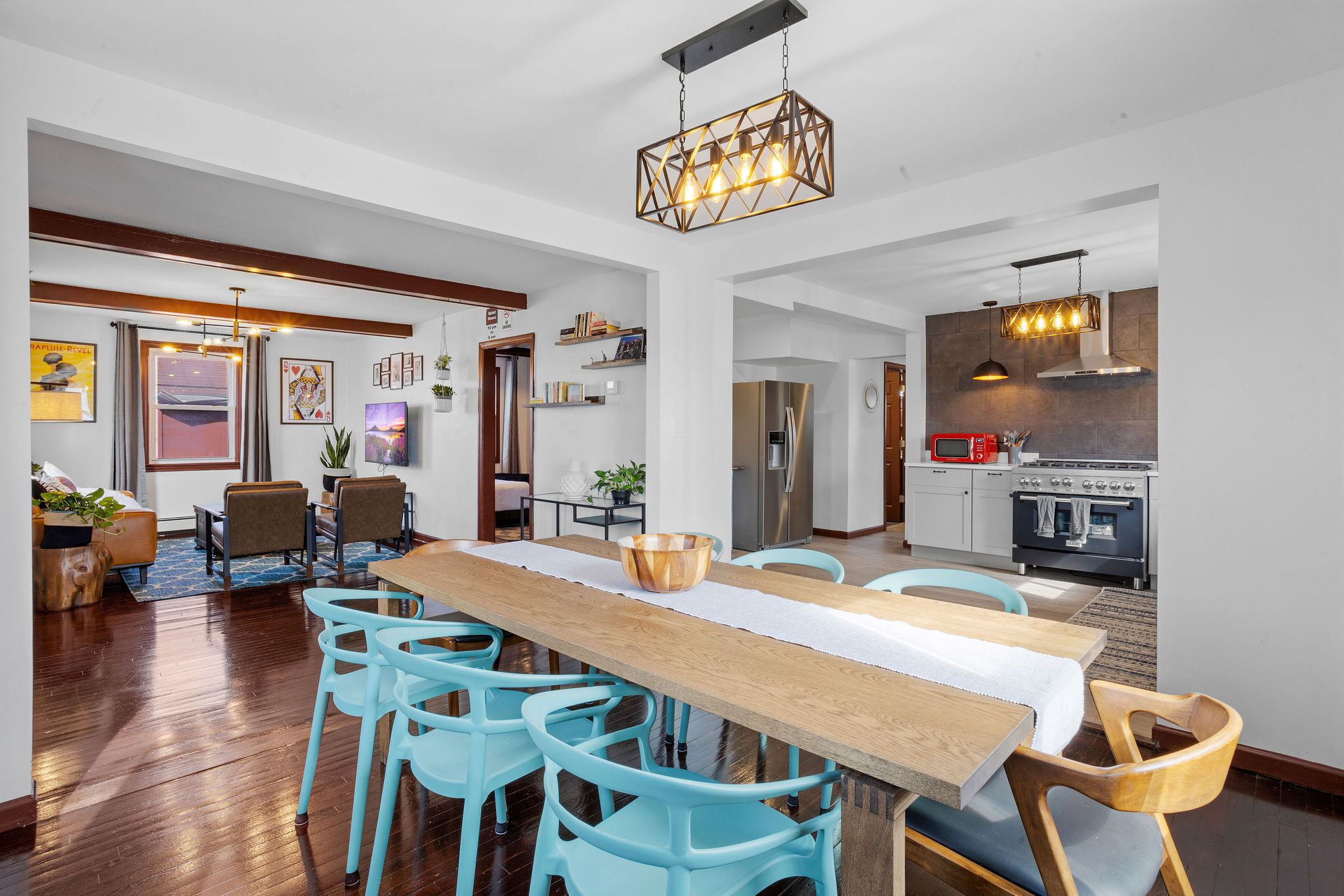 A spacious dining area with a long wooden table and blue chairs, a kitchen with modern appliances, and a hallway with framed artwork.