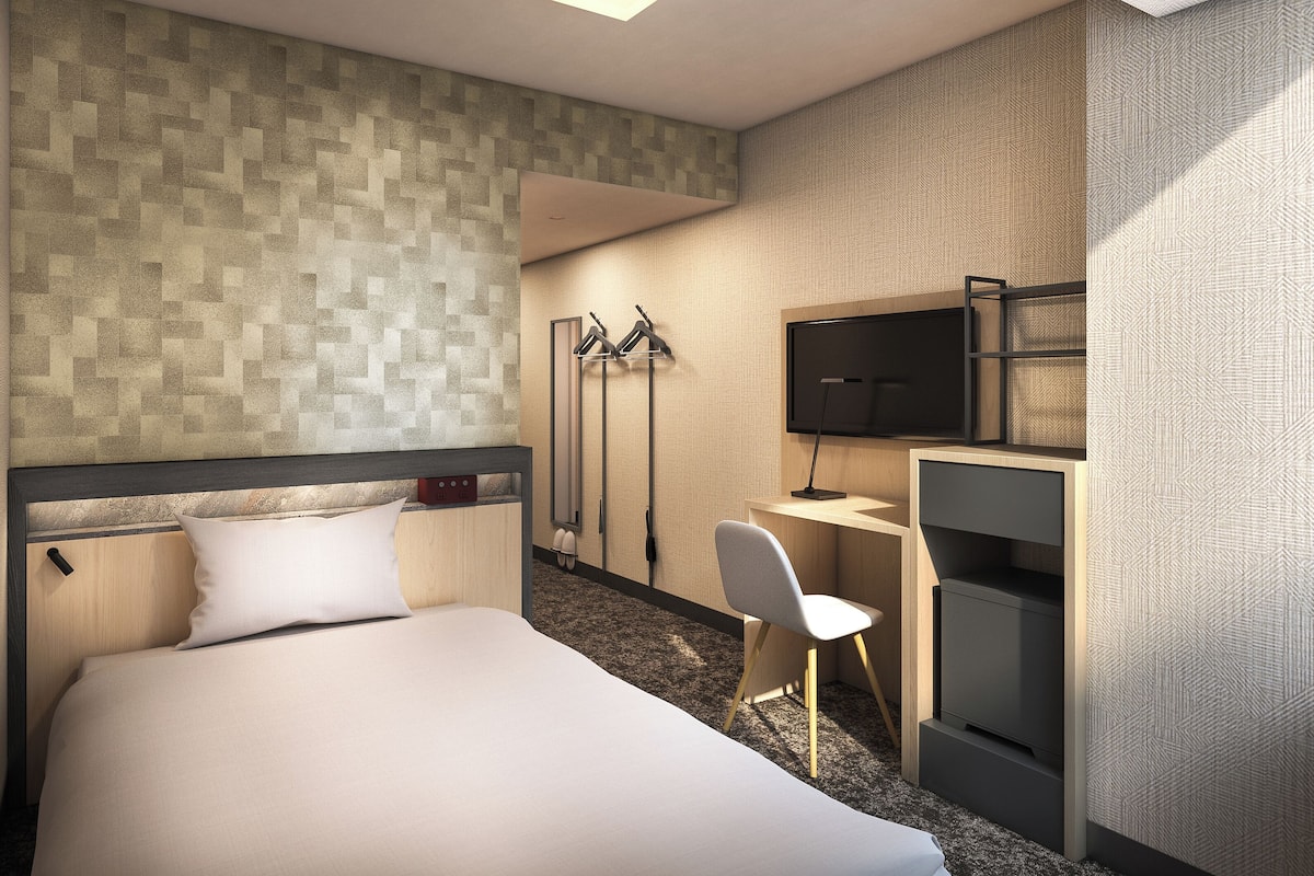 A hotel room with a bed, a desk, a chair, a TV, and a coat rack.