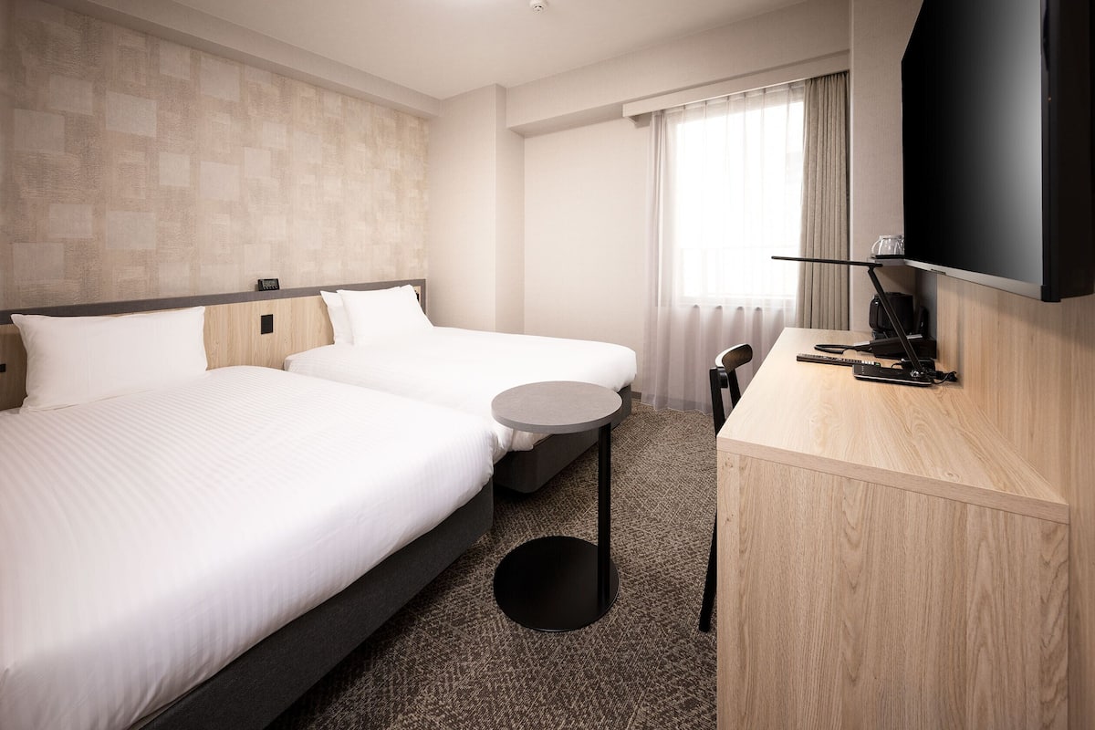 A hotel room with two beds, a small round table, a desk with a flat-screen TV, and a window with curtains.