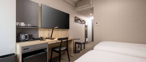 A hotel room with a bed, a desk with a chair, a small refrigerator, a coffee maker, a flat-screen TV, and a wall-mounted air conditioner.