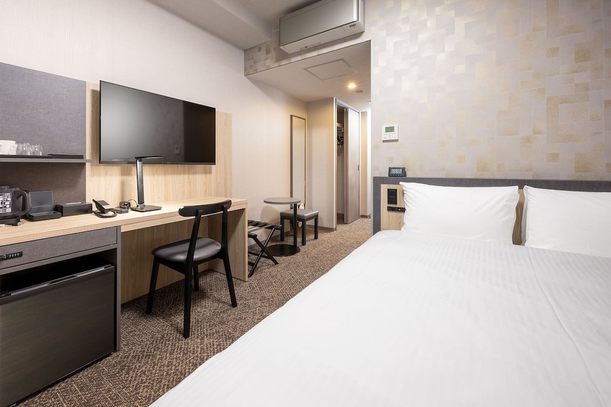 A hotel room with a bed, a desk with a computer, a chair, a small table, a refrigerator, and a TV mounted on the wall.