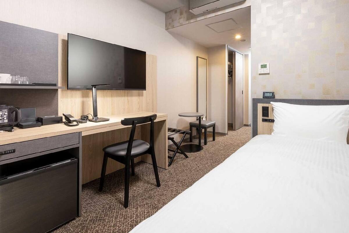 A hotel room with a bed, desk, chair, TV, and a small kitchenette.