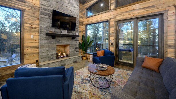 A cozy living room with a stone fireplace, blue sofas, and a large window overlooking a deck. - These Are The Days | Luxury Hochatown Cabin with Hot Tub, Grill & Pet-Friendly Comfort (Hochatown)
