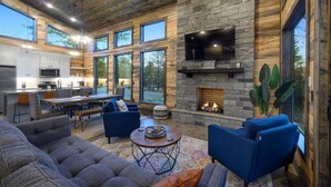 A spacious living area with a stone fireplace, comfortable seating, and large windows offering views of the outdoors. - These Are The Days | Luxury Hochatown Cabin with Hot Tub, Grill & Pet-Friendly Comfort (Hochatown)
