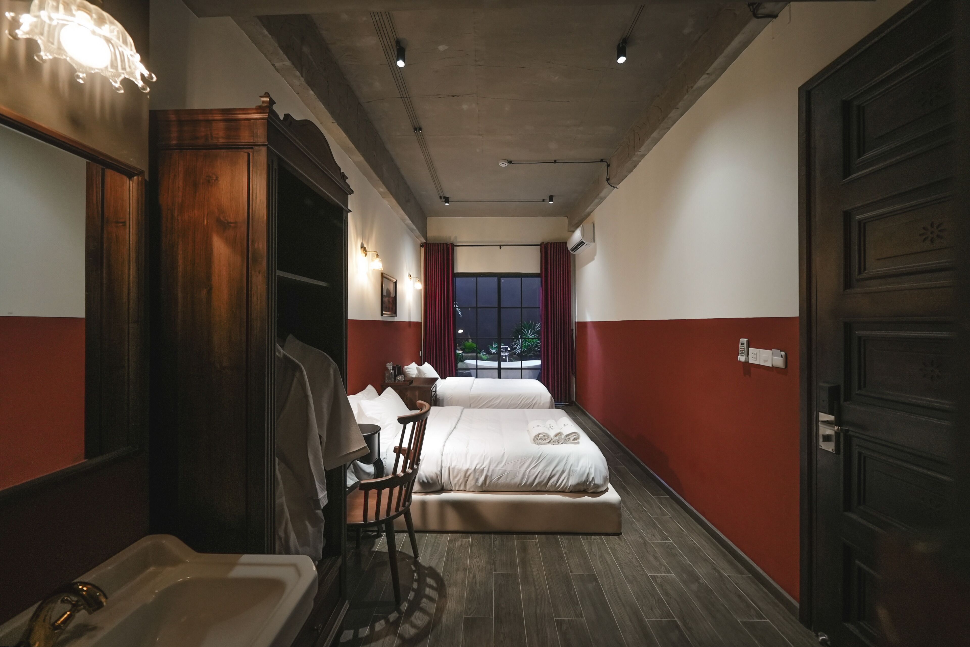 A hotel room with a large bed, a wooden wardrobe, a desk with a chair, and a large window with curtains.