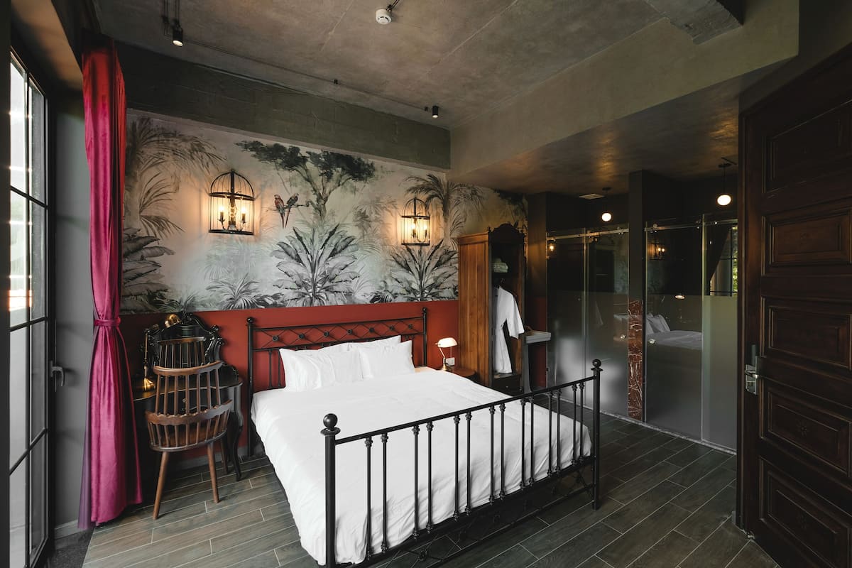 Un dormitorio con cama, mesita de noche, lámpara, estante en la pared y ventana con cortinas.