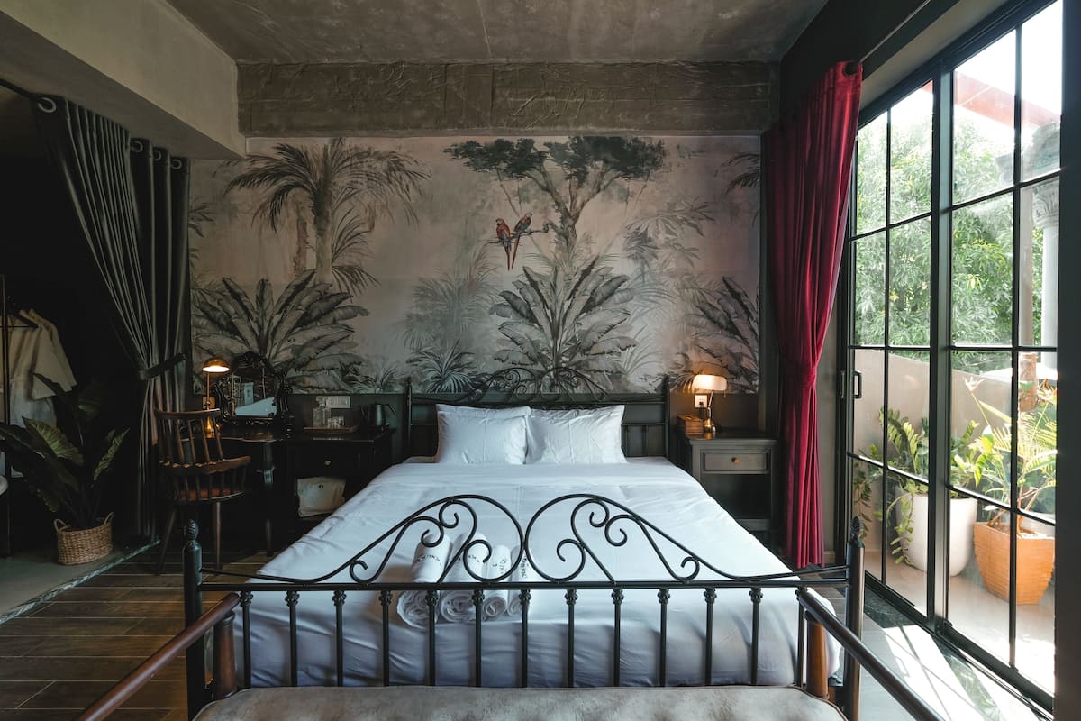 Un dormitorio con una cama grande, un mural de árboles y pájaros, una ventana con cortinas rojas y vistas al verdor exterior.