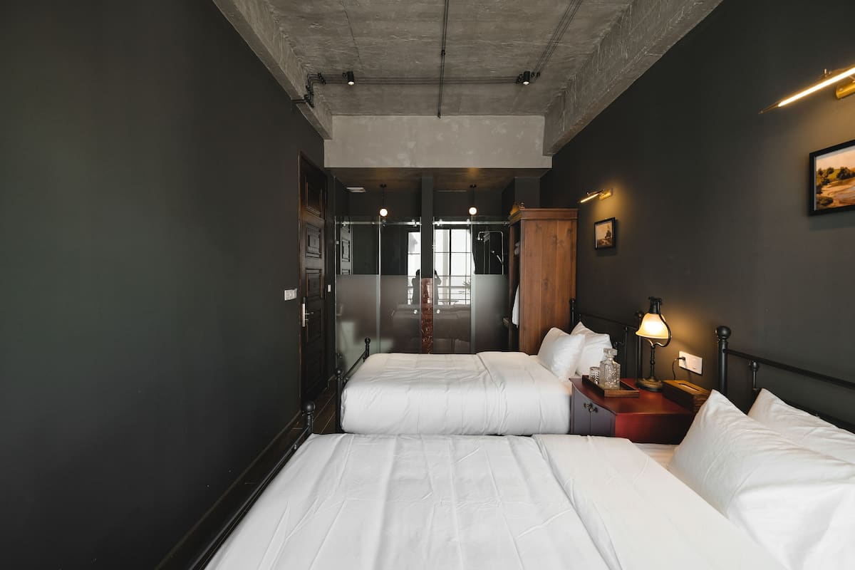 Habitación de hotel con dos camas, un armario de madera, una mesita de noche con lámpara y un cuadro en la pared.