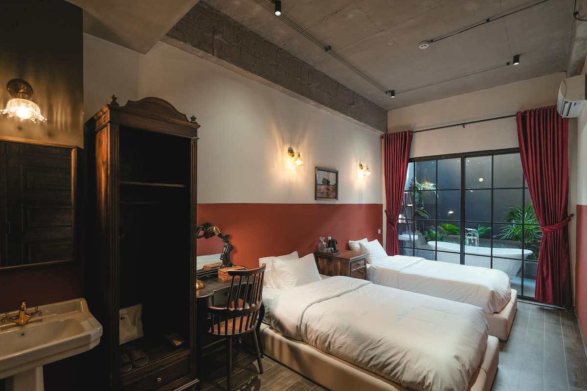 Habitación de hotel con dos camas, un escritorio con silla, un espejo grande y vista al exterior a través de un ventanal.