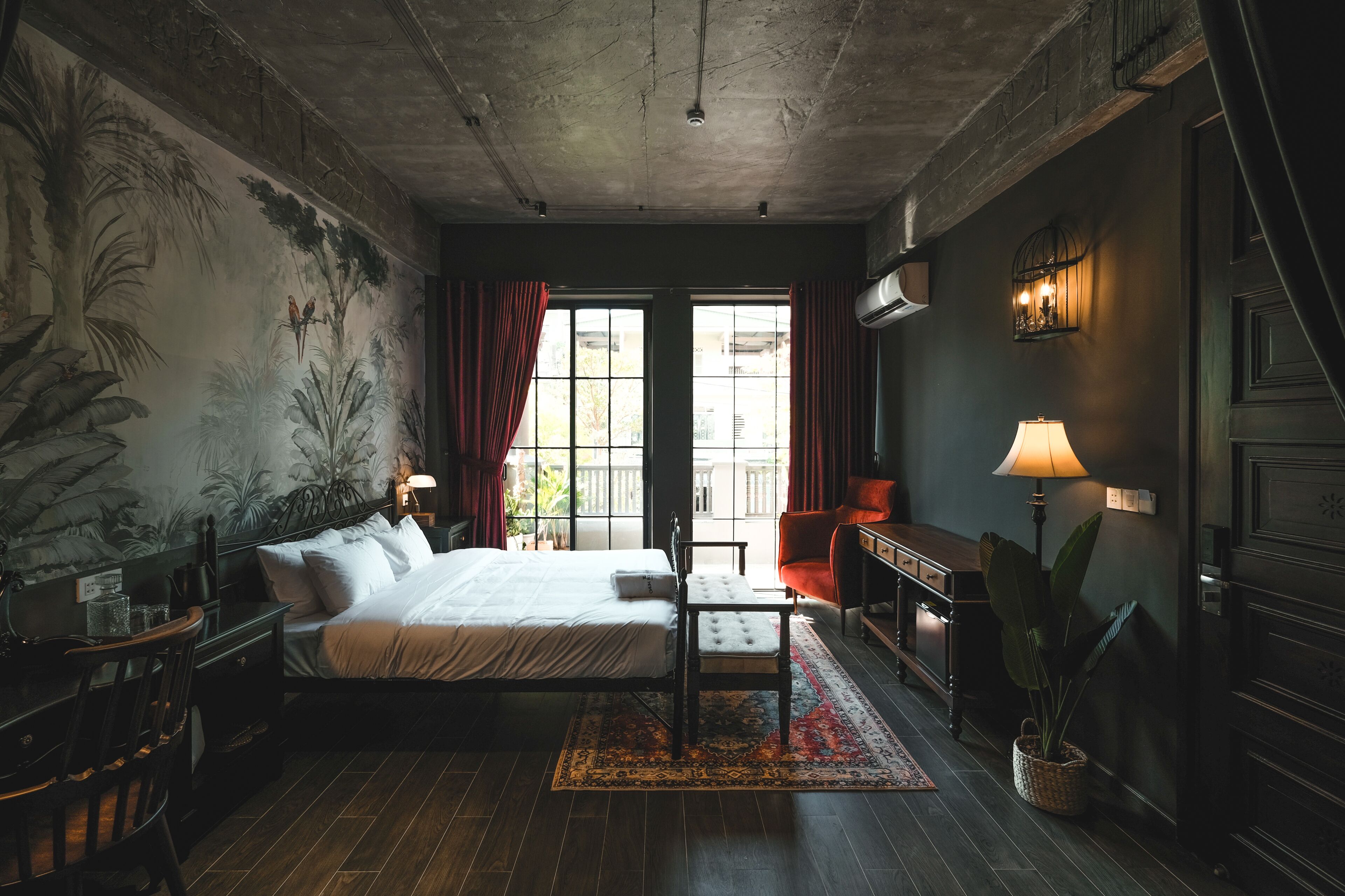 Ein Zimmer mit einem Bett, einem Schreibtisch, einem Stuhl, einer Lampe und einem großen Fenster mit Vorhängen.