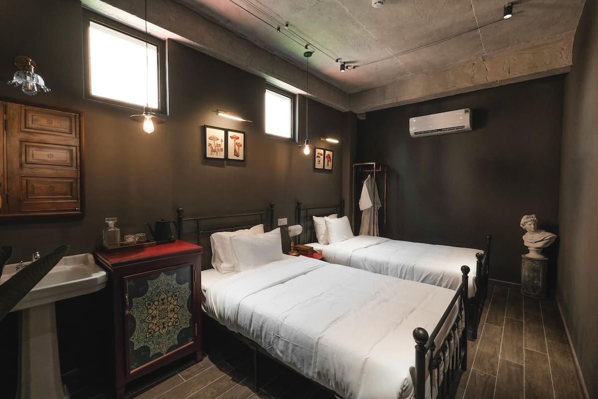 Habitación de hotel con dos camas, un lavamanos, un mueble con azulejos decorativos, cuadros enmarcados en la pared y un equipo de aire acondicionado.