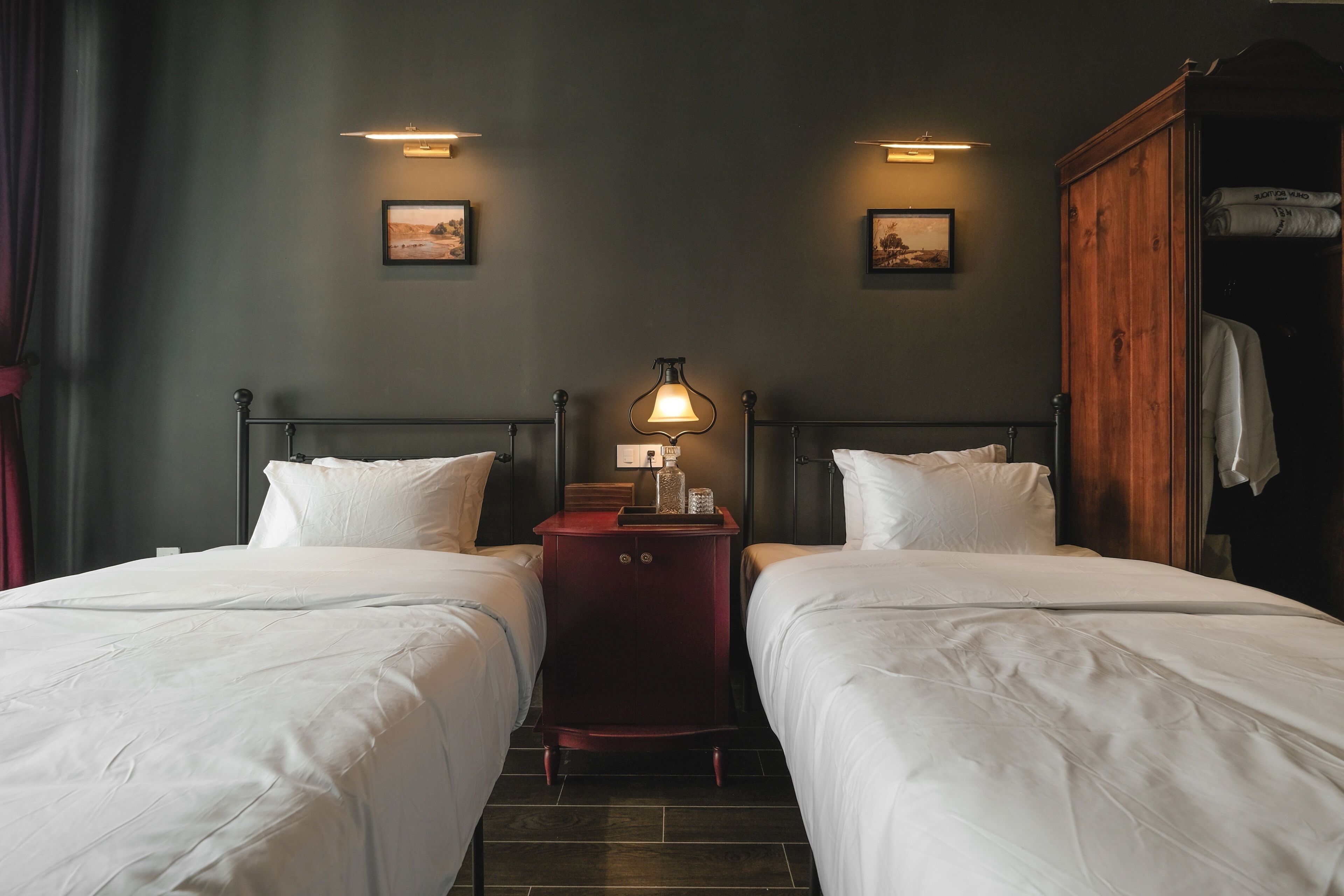 Ein Hotelzimmer mit zwei Betten, einem Holzschrank, einem Nachttisch mit Lampe und gerahmten Bildern an der Wand.
