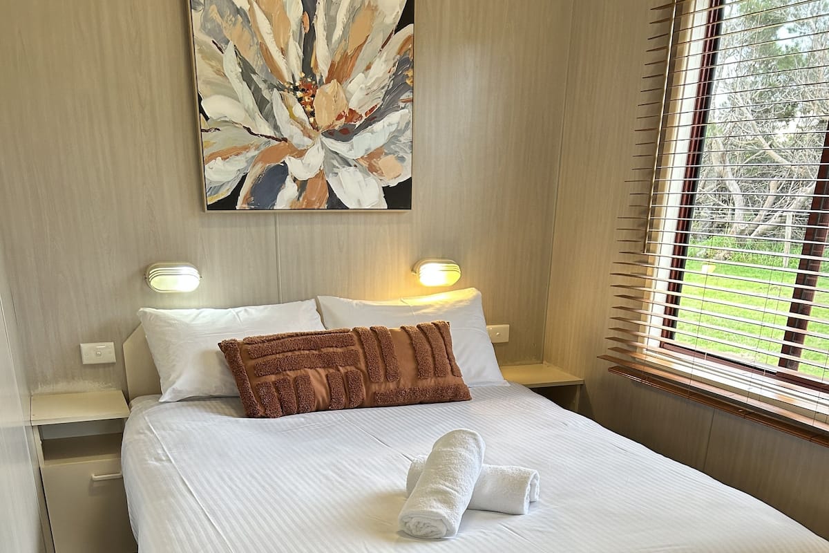 A neatly made bed with white linens and a brown throw pillow, a wall-mounted abstract painting, and a window with blinds.