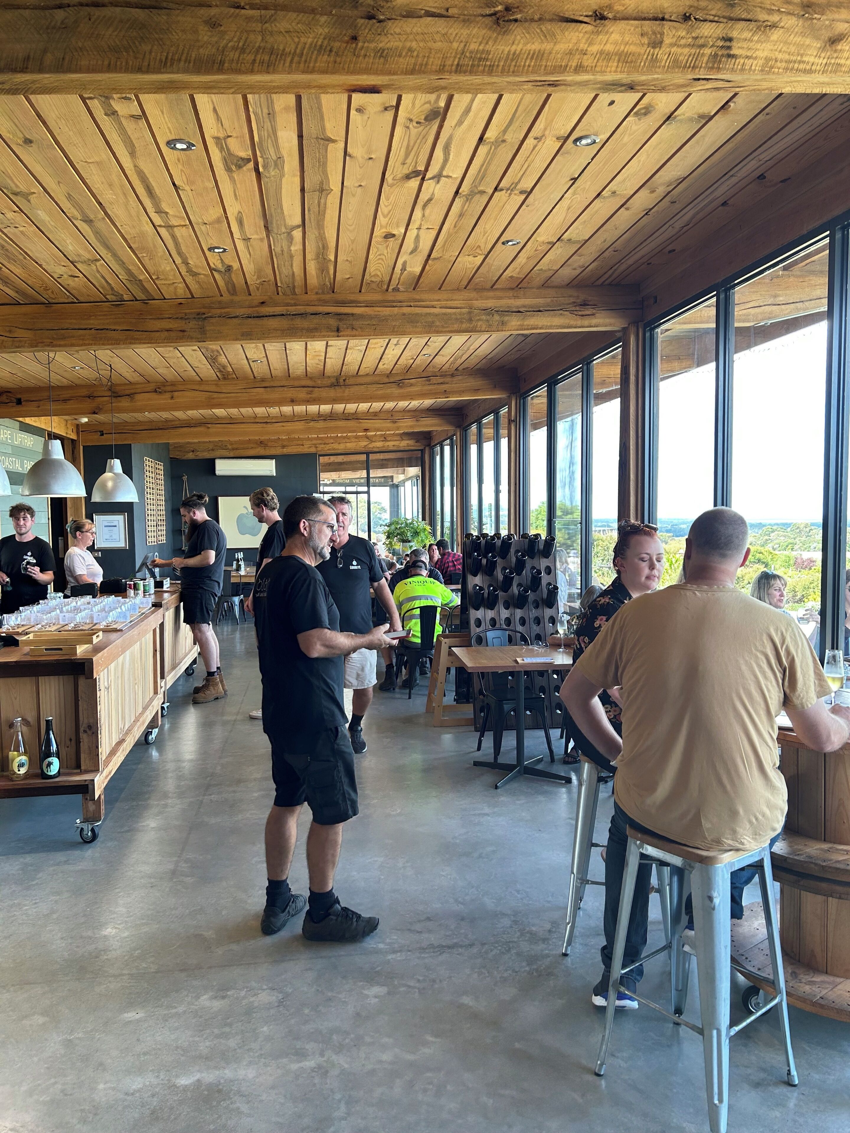 A spacious indoor area with wooden ceiling and large windows, featuring a bar area with bottles and glasses, and a seating area with tables and chairs.