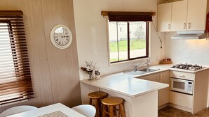 A kitchen with a central island, a dining table with chairs, a clock, and a window with blinds. - Wilsons Prom Holiday Park (Yanakie)