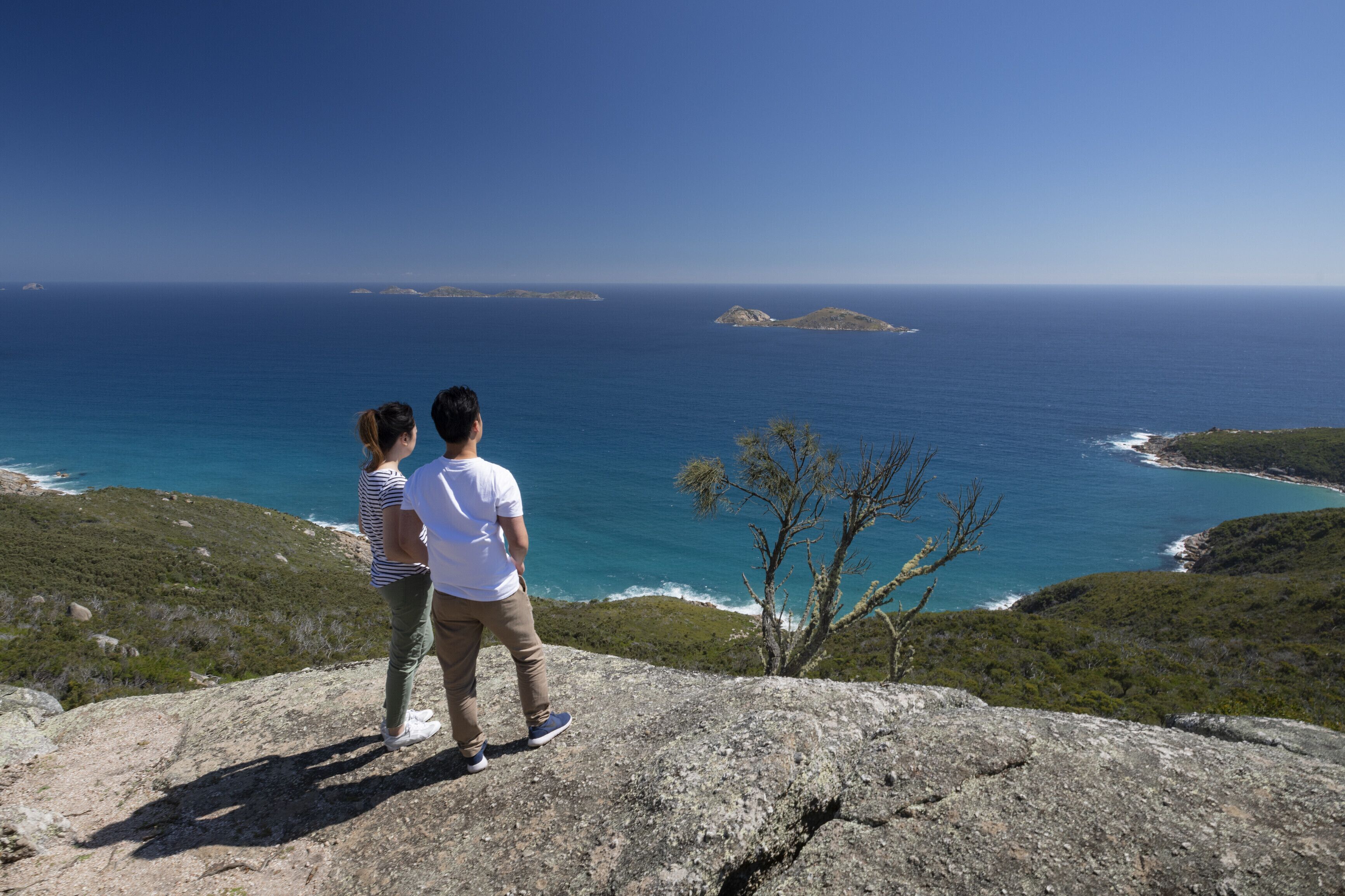 Deux personnes se tiennent sur un promontoire rocheux, d’où elles dominent un paysage côtier avec des eaux d’un bleu limpide et des îles lointaines.
