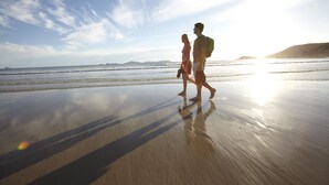 A beach scene with a couple walking on the sand, a clear sky, and distant hills. - Wilsons Prom Holiday Park (Yanakie)