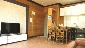 Una sala de estar con una televisión de pantalla plana, piso de madera, un sofá de cuero marrón, una zona de comedor con sillas de mimbre y una cocina con armarios blancos.