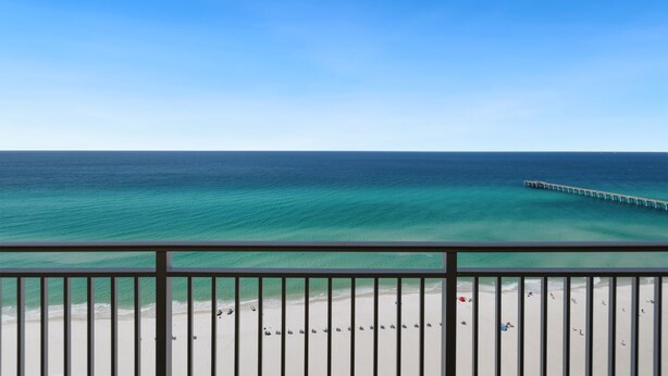 Une vue sur la plage avec une jetée qui s’avance dans l’océan, un ciel dégagé et une balustrade de balcon.