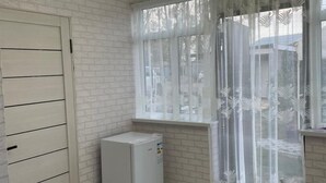 A modern kitchen with white brick walls, a refrigerator, a dining table with chairs, and a window with sheer curtains.