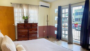 Une chambre à coucher avec un grand lit, une commode en bois, une plante en pot et une fenêtre donnant sur l’extérieur.